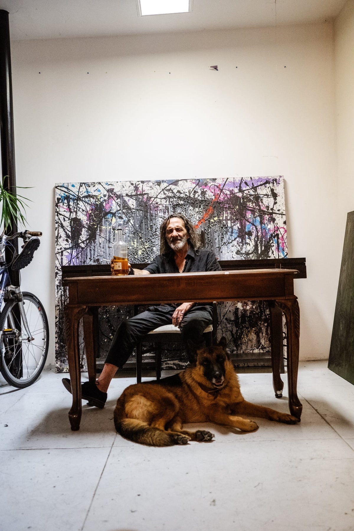 Man seated at wooden desk in art studio with splatter painting and German Shepherd at his feet