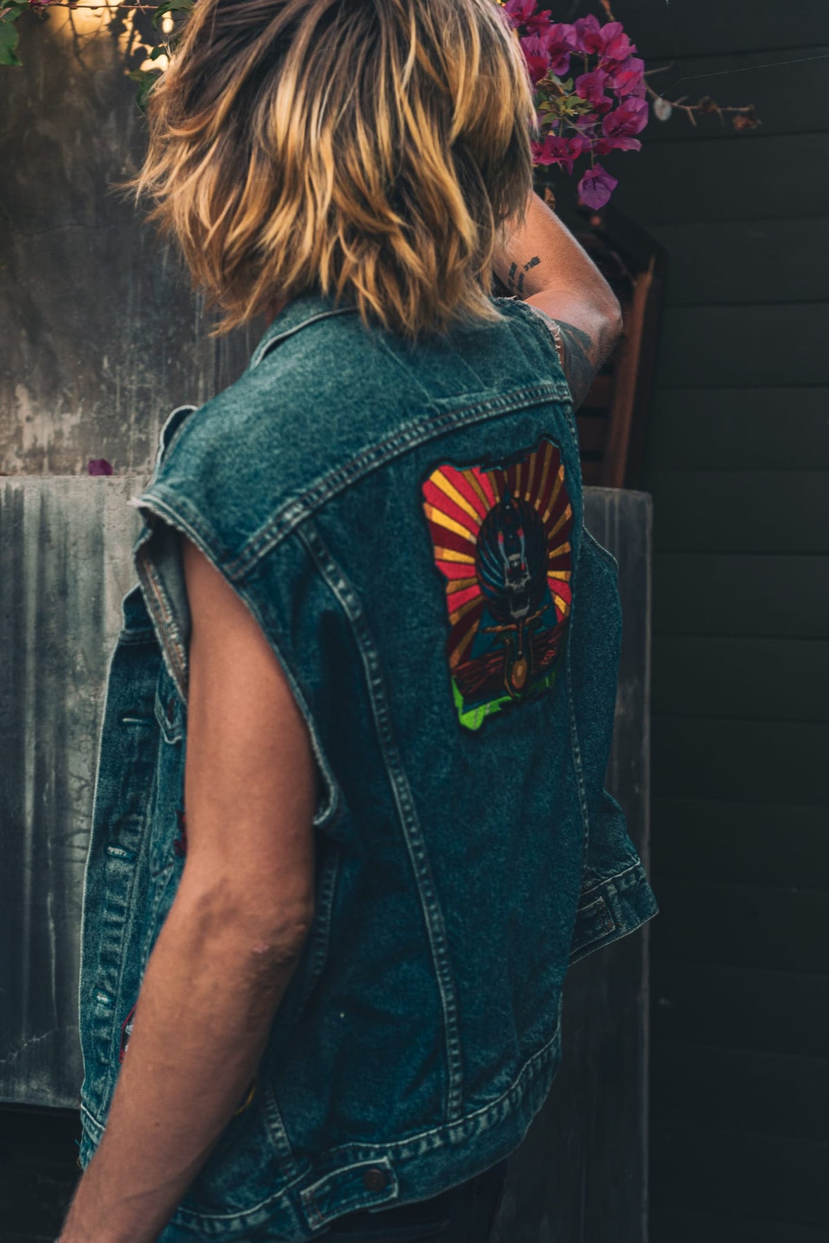 Woman from behind wearing denim vest with psychedelic back patch, bougainvillea in background