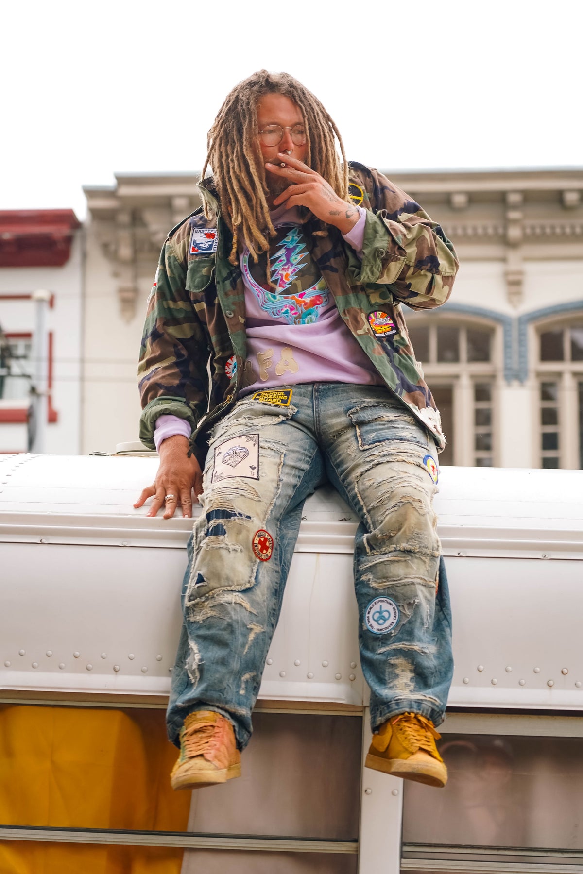 Man with dreadlocks sitting on bus rooftop in camo jacket, patched jeans, and purple hoodie
