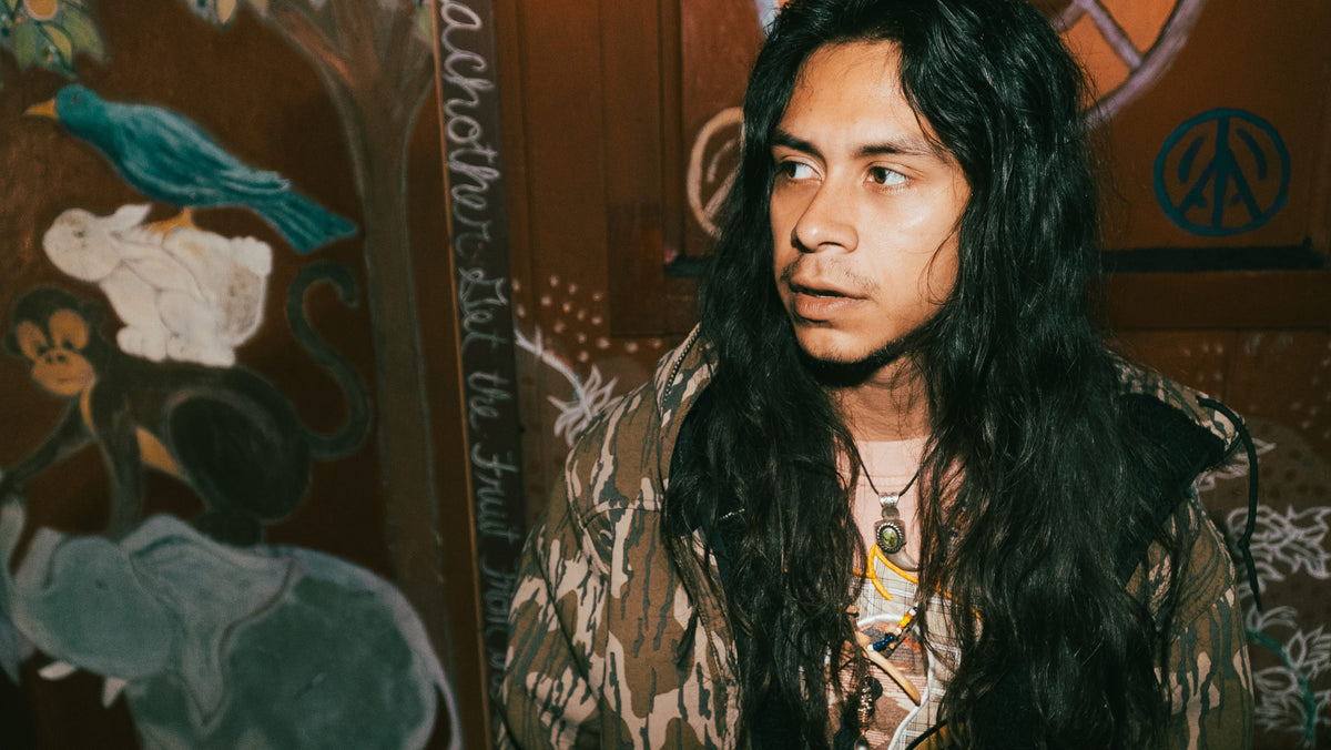 Portrait of man with long black hair in camo jacket next to painted mural with peace sign