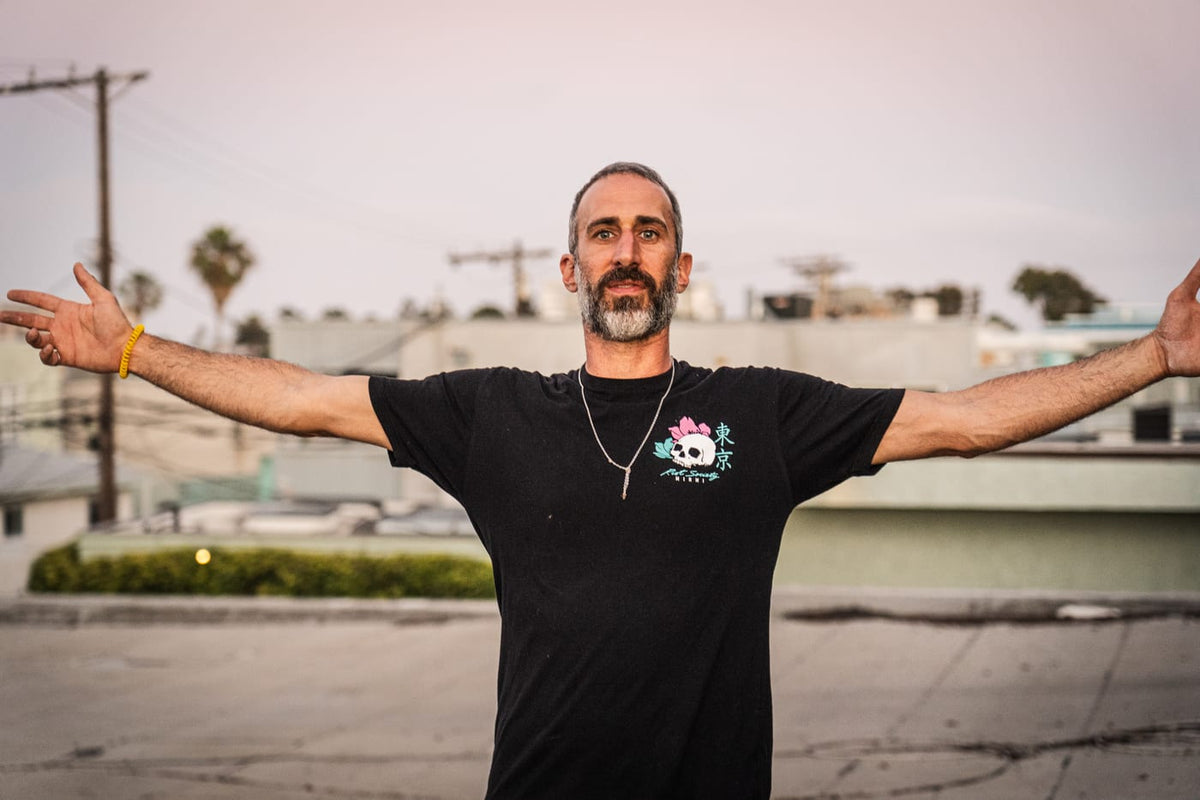 Man in black graphic tee with lotus skull design, arms spread wide on a rooftop at dusk
