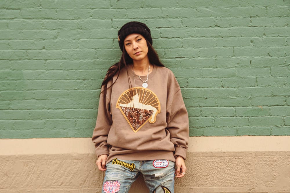 Woman in tan sweatshirt with sunrise cat graphic and patched jeans leaning against green brick wall