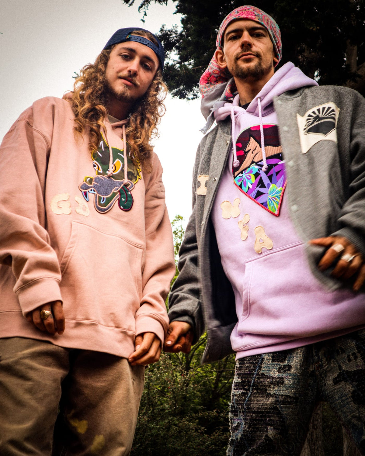 Two men in graphic hoodies shot from low angle against greenery