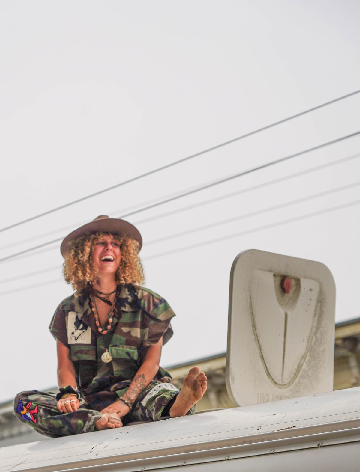 Woman laughing cross-legged on a bus rooftop in camo outfit and wide-brim hat