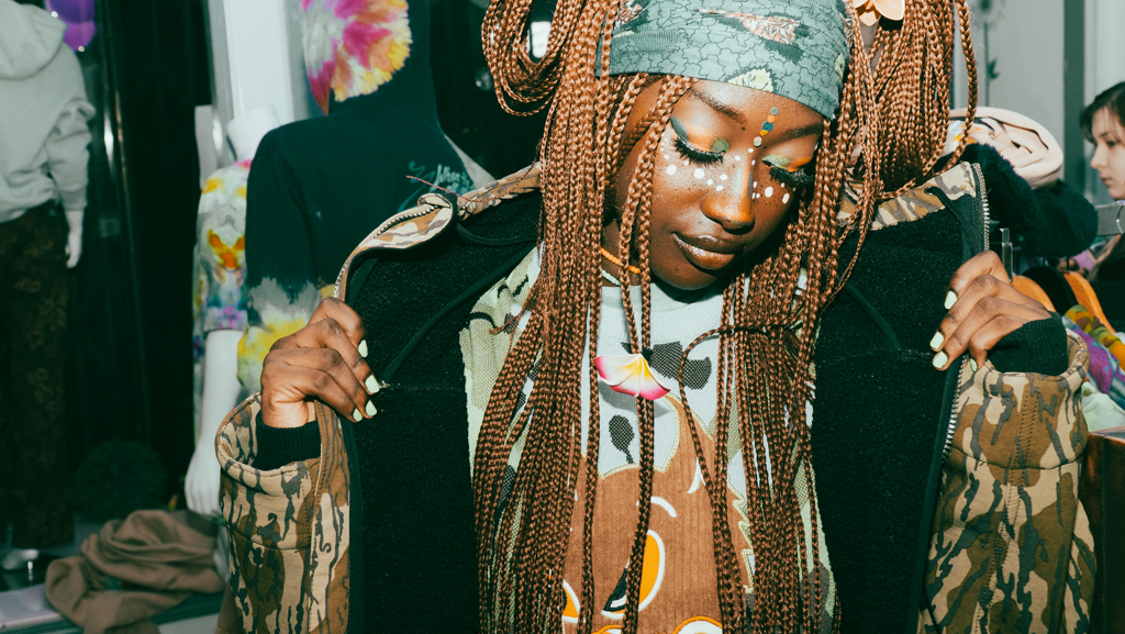 Woman adjusting layered jackets with braids and festival face paint in warm-toned indoor setting